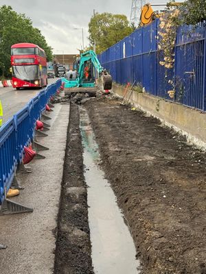 Excavator removing the existing footway beside live traffic under barriered traffic management on River Road, Barking
